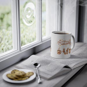 May include: A white ceramic mug featuring the word Autumn in brown script, accompanied by illustrations of a steaming mug, patterned socks, and a candle. The mug sits on an open book next to a small plate of cookies and a silver spoon.