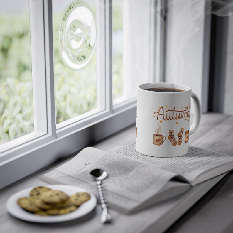 May include: A white ceramic mug featuring the word Autumn in brown script, accompanied by illustrations of a steaming mug, patterned socks, and a candle. The mug sits on an open book next to a small plate of cookies and a silver spoon.