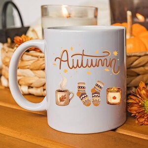 May include: A white ceramic mug featuring the word Autumn in brown cursive script, accompanied by illustrations of a steaming mug of coffee with a leaf, a pair of patterned knit socks, and a small candle with a pumpkin graphic on the front.