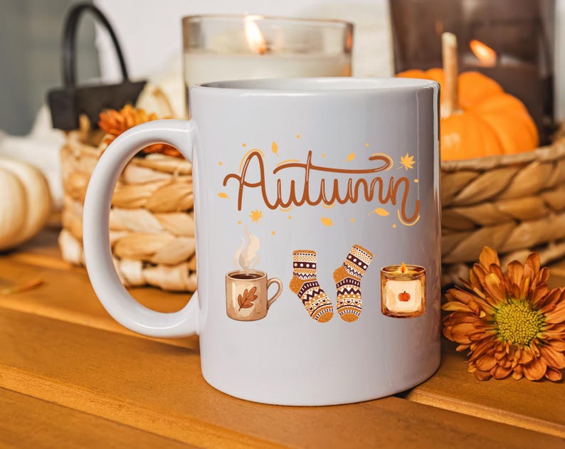 May include: A white ceramic mug featuring the word Autumn in brown cursive script, accompanied by illustrations of a steaming mug of coffee with a leaf, a pair of patterned knit socks, and a small candle with a pumpkin graphic on the front.