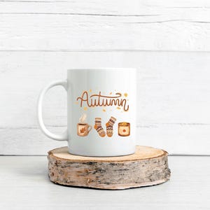 May include: A white ceramic mug featuring the word Autumn in brown script, accompanied by illustrations of a steaming mug, a pair of patterned knit socks, and a lit candle, all rendered in warm orange, brown, and cream color tones.