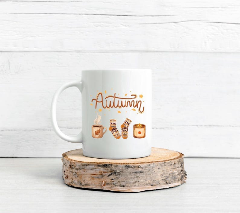 May include: A white ceramic mug featuring the word Autumn in brown script, accompanied by illustrations of a steaming mug, a pair of patterned knit socks, and a lit candle, all rendered in warm orange, brown, and cream color tones.