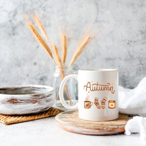 May include: A white ceramic mug featuring the word Autumn in brown script, accompanied by illustrations of a steaming mug, a pair of patterned socks, and a lit candle. The design is centered on the front of the mug, which has a handle on the left side.