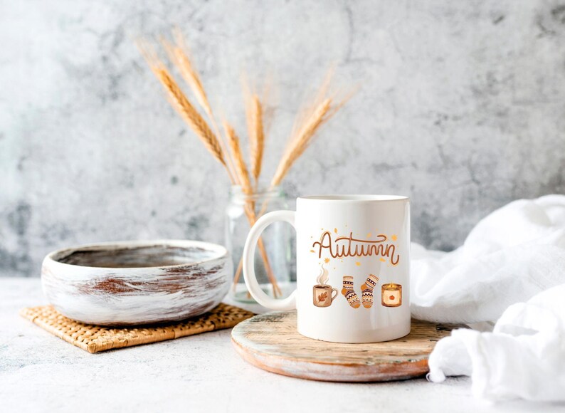May include: A white ceramic mug featuring the word Autumn in brown script, accompanied by illustrations of a steaming mug, a pair of patterned socks, and a lit candle. The design is centered on the front of the mug, which has a handle on the left side.