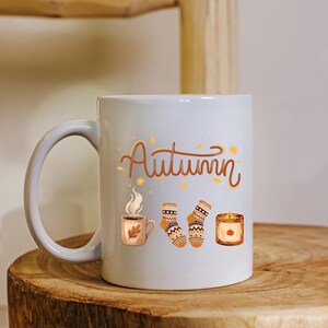 May include: A white ceramic mug featuring the brown cursive text Autumn, accompanied by illustrations of a steaming mug with a leaf, a pair of patterned knit socks, and a lit candle in a glass jar, all decorated with small scattered orange leaves.