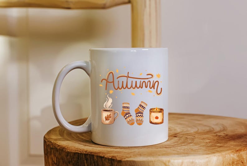 May include: A white ceramic mug featuring the brown cursive text Autumn, accompanied by illustrations of a steaming mug with a leaf, a pair of patterned knit socks, and a lit candle in a glass jar, all decorated with small scattered orange leaves.