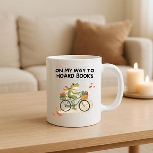 Könnte beinhalten: Ein weißer Keramikbecher mit einer Grafik eines grünen Frosches, der auf einem Fahrrad fährt und Bücher in einem Korb transportiert. Der Text ON MY WAY TO HOARD BOOKS steht über dem Frosch, ergänzt durch kleine orangefarbene und braune Herbstblätter.