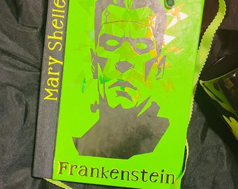 May include: A green book with a silver silhouette of Frankenstein's monster on the cover. The book title "Frankenstein" is written in gold lettering on the bottom of the cover. The author's name, "Mary Shelley", is written in gold lettering on the spine of the book.