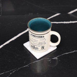 May include: A white ceramic mug with a teal blue interior and a black and white illustration of the state of Pennsylvania with the text "Pennsylvania" and a list of cities. The mug is sitting on a white square coaster.