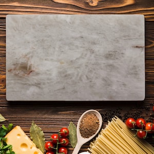 May include: A rectangular light gray marble cutting board with natural veining patterns. The surface is smooth and polished, featuring subtle white and soft brown mineral streaks throughout the stone slab, providing a neutral backdrop.