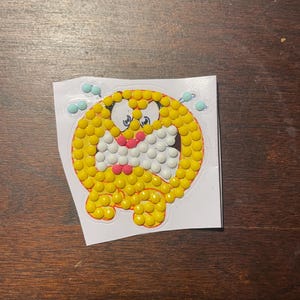May include: A sticker featuring a cartoon character with a yellow body, white mouth, and pink nose. The character has large eyes and is surrounded by small blue shapes. The sticker is on a white backing.