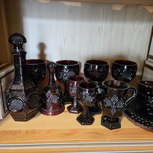 May include: A collection of dark red pressed glass items including a decanter with a stopper, a bell, several stemmed goblets, a handled mug, and a small bowl, all featuring a repeating geometric pattern with circular and diamond motifs.