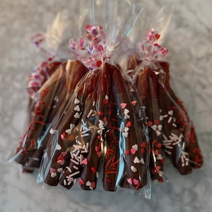 May include: Chocolate-covered pretzel sticks decorated with red, white, and pink heart-shaped sprinkles and red sprinkles. Each is individually wrapped in clear cellophane and tied with a pink and red heart-patterned ribbon.