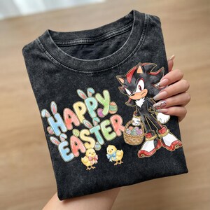 May include: Black t-shirt with a colorful "Happy Easter" graphic, bunny ears, and two yellow chicks. The shirt features a cartoon character holding a basket of Easter eggs. The t-shirt is made of a soft, comfortable material.