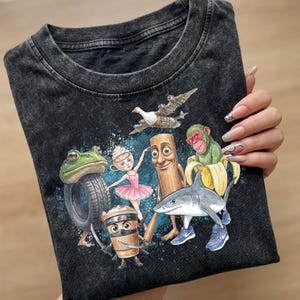 Może przedstawiać: Ciemnoszara koszulka z fantazyjną ilustracją różnych postaci, w tym żaby z oponą, baletnicy, filiżanki kawy ninja, rekina w trampkach, małpy i drewnianej figurki. Projekt umieszczono na niebieskim tle.