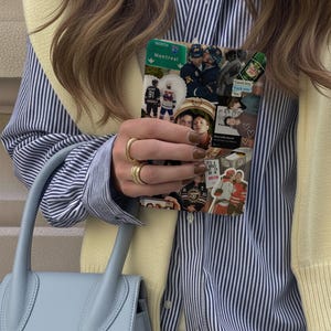 K&ouml;nnte beinhalten: Eine hellblaue Handtasche mit gebogenem Griff wird in der N&auml;he eines gestreiften blau-wei&szlig;en Hemdes gehalten. Eine Handyh&uuml;lle mit einer Collage von Bildern, darunter Eishockeyspieler und Text, wird vor dem Hemd gehalten. Goldringe schm&uuml;cken die Finger.
