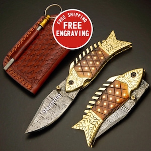 May include: Two fish-shaped folding knives with ornate gold and wood handles. The blades have a unique pattern and the word "Surprise" is etched on one. A brown leather case with a sharpening rod is also included.