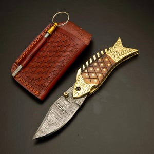 May include: A decorative folding knife in the shape of a fish, with a patterned blade and a brass and wood handle. The knife is accompanied by a brown leather sheath and a sharpening rod. The blade has a unique, wavy pattern.
