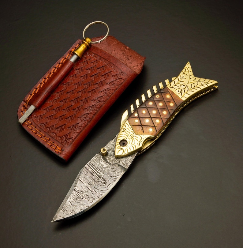 May include: A decorative folding knife in the shape of a fish, with a patterned blade and a brass and wood handle. The knife is accompanied by a brown leather sheath and a sharpening rod. The blade has a unique, wavy pattern.