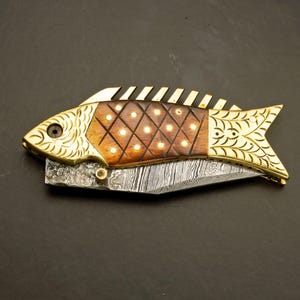 May include: A decorative folding knife in the shape of a fish. The knife features a patterned blade, a wooden body with inlaid dots, and gold-colored metal accents. The fish knife is a unique collectible.