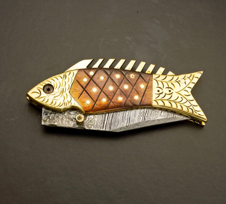 May include: A decorative folding knife in the shape of a fish. The knife features a patterned blade, a wooden body with inlaid dots, and gold-colored metal accents. The fish knife is a unique collectible.