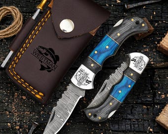 May include: A folding pocket knife with a Damascus steel blade and a blue and black wood handle, featuring the name GEORGE engraved on the bolster. It is paired with a brown leather sheath embossed with the name WESTMAN and a fish graphic.