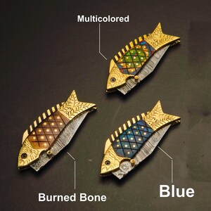 May include: Three decorative fish-shaped knives with gold-colored accents. One knife has a multicolored design, another has a burned bone design, and the third has a blue design. The knives are on a dark background.
