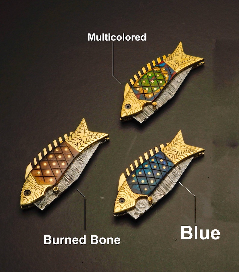 May include: Three decorative fish-shaped knives with gold-colored accents. One knife has a multicolored design, another has a burned bone design, and the third has a blue design. The knives are on a dark background.