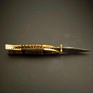 May include: A decorative folding knife with a gold-colored handle and a patterned blade. The handle features intricate designs with colorful accents. The blade is pointed and has a textured appearance. The knife is displayed on a dark surface.