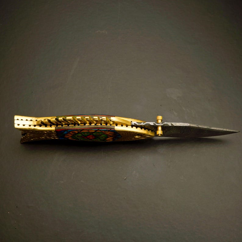 May include: A decorative folding knife with a gold-colored handle and a patterned blade. The handle features intricate designs with colorful accents. The blade is pointed and has a textured appearance. The knife is displayed on a dark surface.