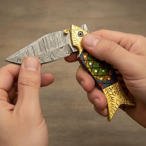 May include: A decorative folding knife with a fish-shaped handle. The blade has a patterned design. The handle is gold, with colorful geometric inlays of green, blue, and red. The knife is held open, displaying the blade.