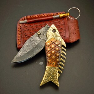 May include: A decorative folding knife with a fish-shaped handle. The blade is open, revealing a patterned steel surface. The handle is made of wood and brass, with a leather sheath and sharpening tool in the background.