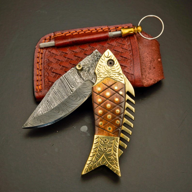 May include: A decorative folding knife with a fish-shaped handle. The blade is open, revealing a patterned steel surface. The handle is made of wood and brass, with a leather sheath and sharpening tool in the background.