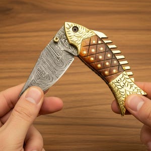 May include: A decorative folding knife in the shape of a fish. The blade has a unique patterned design. The handle is made of wood and brass, with a fish scale pattern. The knife is held open, displaying the blade.