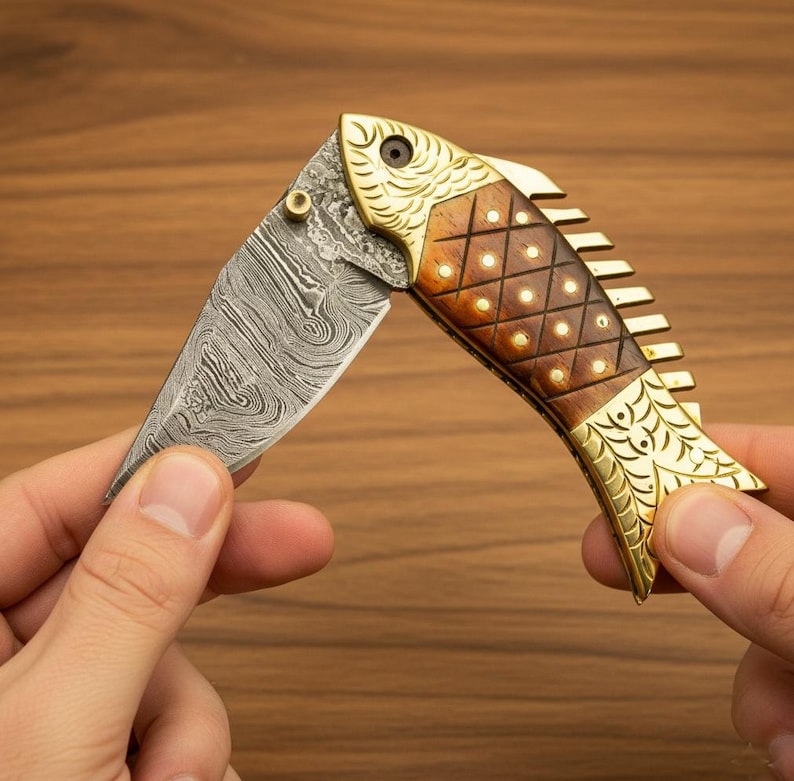 May include: A decorative folding knife in the shape of a fish. The blade has a unique patterned design. The handle is made of wood and brass, with a fish scale pattern. The knife is held open, displaying the blade.
