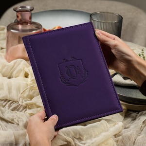 May include: A rectangular purple menu cover featuring a debossed crest logo in the center. The cover has stitched edges and a smooth, matte finish. The design is simple and functional, providing a structured surface for holding documents or menus.