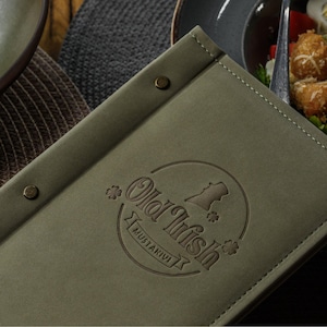 Può includere: Una copertina per menu rettangolare color verde oliva con un logo circolare impresso recante il testo Old Irish e Mustakivi, accentuato da piccole icone a forma di trifoglio e due rivetti metallici sul bordo sinistro, con bordi cuciti.