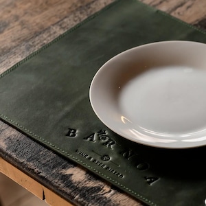 May include: A rectangular dark green leather placemat with visible stitching along the edges. The surface features an embossed logo that reads BAR NOA WINE & BEER BISTRO. A plain white ceramic plate is positioned on the right side of the placemat.