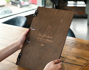 May include: A dark brown wooden menu cover with black leather hinges and metal rings. The cover features an engraved lighthouse icon and the text "le Bistrot Hôtel du Phare BELLE-ÎLE-EN-MER" centered on the front surface of the rectangular board.