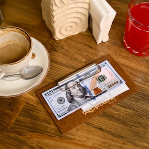 May include: A brown leather billfold with a metal clip holding a $100 bill. The billfold is on a wooden table with a cup of coffee and a glass of red juice.