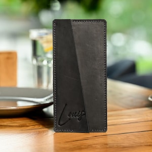 May include: A black leather wallet with a diagonal design and the name "Louis" embossed on the front. The rectangular wallet is hand-stitched. The background is blurred, suggesting a restaurant setting.