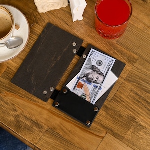 May include: A dark brown wooden bill holder with a black leather pocket, containing a £100 bill and a white receipt. The holder is open, revealing the contents. The scene is set on a wooden table, with a cup of coffee and a red drink in the background.