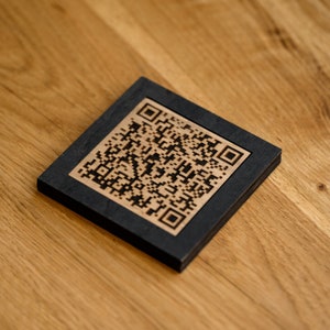Op de afbeelding: Een vierkante houten onderzetter met een QR-code ontwerp. De onderzetter heeft een zwart kader met een lichtbruin middenstuk met de QR-code. Hij ligt op een houten oppervlak.