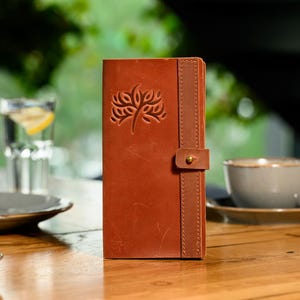 May include: A brown leather journal with an embossed tree design on the front. It features a vertical strap with a brass button closure. The journal is on a wooden table. The journal measures approximately 20 cm in height.