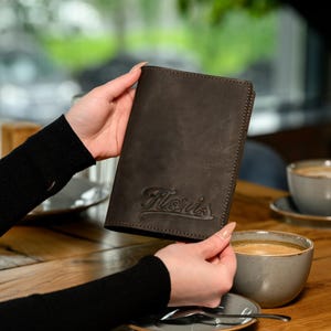 Op de afbeelding: Een donkerbruine leren portemonnee met het woord "Floris" in reliëf in een scriptlettertype. De portemonnee is rechthoekig en lijkt gesloten. De achtergrond omvat een houten tafel en een kopje koffie.
