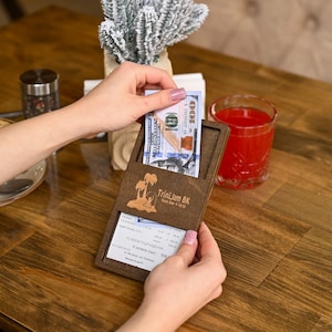 May include: A wooden bill holder with the text "TriniJam BK Roti Bar & Grill" and a banknote inside. The bill holder is on a wooden table with a glass of red liquid.