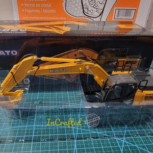 Könnte beinhalten: Ein gelbes und schwarzes Modell einer hydraulischen Baggers der Marke Kato HD820 mit einer 90-Grad-Articulation. Das Modell wird in einem durchsichtigen Kunststoffgehäuse auf einer grünen Gitteroberfläche präsentiert.