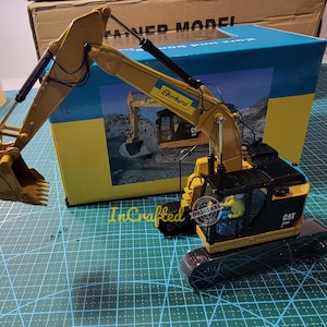 Op de afbeelding: Een gele en zwarte speelgoedgraafmachine met een zwarte cabine en een gele arm. De graafmachine staat op een groene mat en heeft het woord "CAT" aan de zijkant. De graafmachine is een model op schaal 1:20.