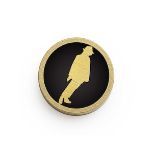 May include: A circular, gold-coloured pin with a black centre. It features a gold silhouette of a person in a hat, in a walking pose. The pin has a textured, wood-like border.