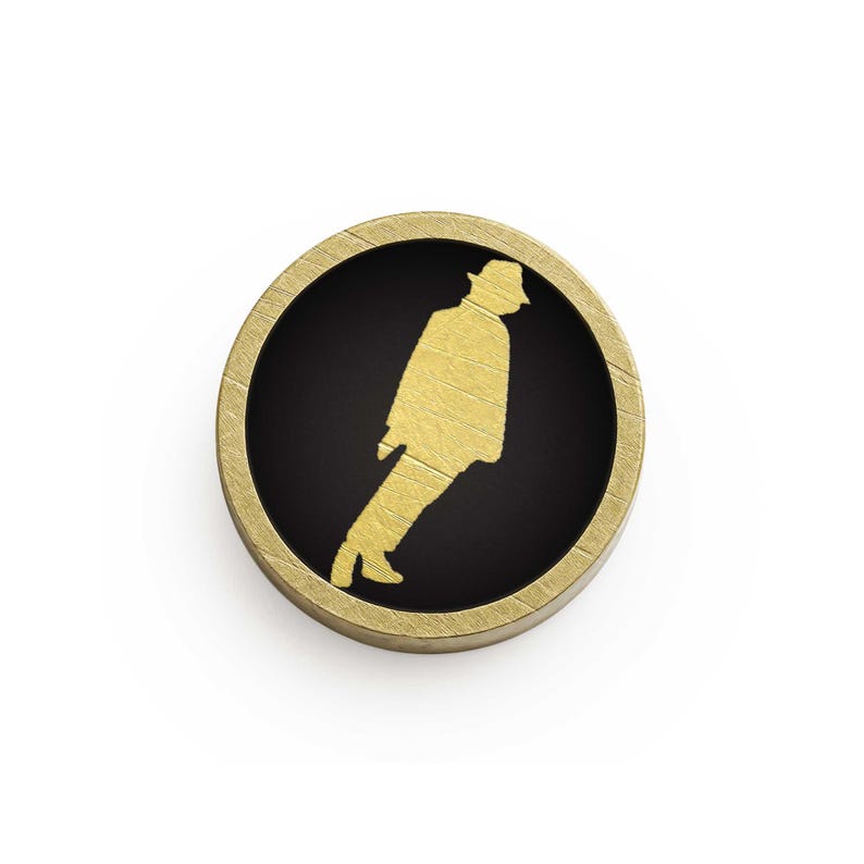 May include: A circular, gold-coloured pin with a black centre. It features a gold silhouette of a person in a hat, in a walking pose. The pin has a textured, wood-like border.
